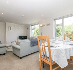 A living room with sofas a dining table set with bowls and glasses and large windows showing trees at Godrevy in Porthcurno