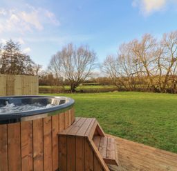 A wooden hot tub with steps on a deck in a grassy yard with leafless trees and a wooden fence at Otter's View in South Luffenham near Edith Weston