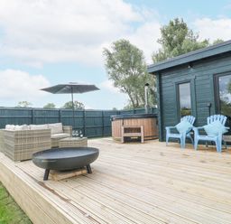 An outdoor wooden deck with a wicker sofa, umbrella, fire pit, hot tub, and blue chairs at Otters View South Luffenham near Edith Weston