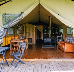 An open tent with wooden flooring containing dining table with chairs a wood stove a leather sofa and shelves with kitchen items at Oak Stockland near Dalwood