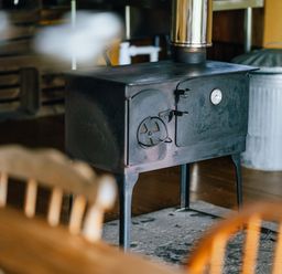 A black wood stove with a thermometer in a room with wooden chairs and a metal trash can