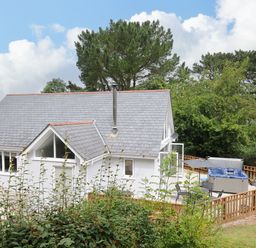 A white house with a gray roof and a fenced deck with a hot tub and outdoor chairs surrounded by trees and bushes at Otters Leap in Bideford