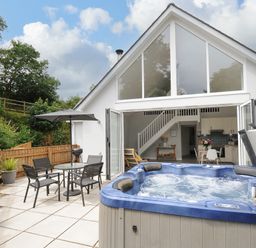 An outdoor patio area with a hot tub table and chairs and an umbrella outside a house with large windows and open doors at Otters Leap in Bideford