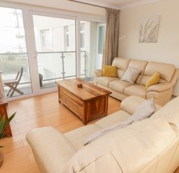 A living room with two beige leather sofas a wooden coffee table a TV on a wooden stand sliding glass doors leading to a balcony with chairs and plants at 54 Croft Court in Tenby