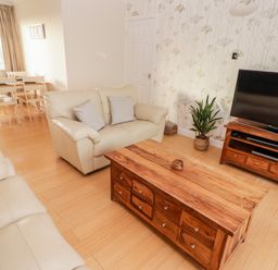 A living room with two beige sofas a wooden coffee table a wooden TV stand with a flat screen TV and a dining table with chairs by a window at 54 Croft Court in Tenby