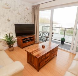 A living room with a wooden coffee table two beige sofas a television on a wooden stand a potted plant and a glass sliding door leading to a balcony at 54 Croft Court in Tenby