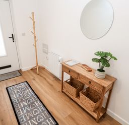 A hallway with a white door a wooden coat rack a wooden console table with baskets a plant and a circular mirror at Puffins Rest in Goldsithney