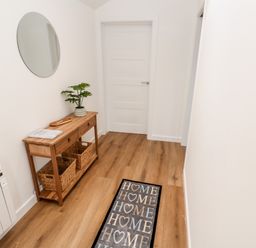 A hallway with wooden floor a wooden shelf with baskets a round mirror and a rug with home text at Puffins Rest in Goldsithney