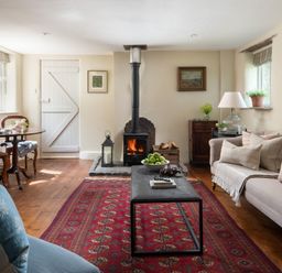 A living room with a wood stove a red patterned rug a beige sofa and a small dining table at Hawthorn Cottage at Collihole in Chagford