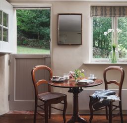 A small dining area with a round wooden table and two wooden chairs near a door and window with flowers on the table at Hawthorn Cottage at Collihole in Chagford