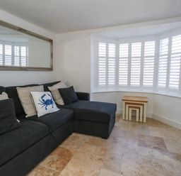 A living room with a dark sectional sofa and cushions near a window with white shutters and nested wooden tables at Belvedere Cottage in Weymouth