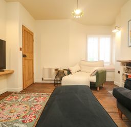 A living room with a patterned rug a wooden door a white cushioned chair a window with shutters a mounted television and a stone fireplace at Josie in Pentewan