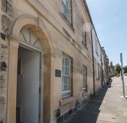 Stone building with arched doorway and windows along a sidewalk on a street with trees and cars at Eider Cottage in Warkworth