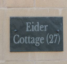 A black sign with white lettering reading Eider Cottage 27 mounted on a stone wall at Eider Cottage in Warkworth