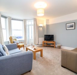 A living room with a blue sofa, a brown chair, a small wooden table with a bottle and glasses, a television on a wooden stand, and framed picture on the wall at 11 The Craighurst in Southwold
