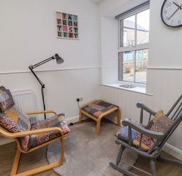 A seating area with a wooden chair, gray rocking chair, footstool, floor lamp, wall clock, and window at 2A Front Street Embleton