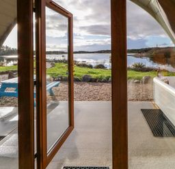 An open glass door looking out to a patio with a blue picnic table and a hot tub beside a lake at The Wheelhouse Pod No 1 in Burtonport County Donegal
