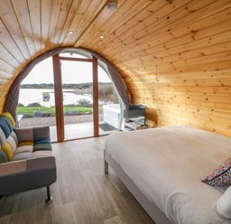 An arched wooden room with a bed, sofa, small TV, and glass door overlooking a lake at The Wheelhouse Pod No 1 Burtonport County Donegal