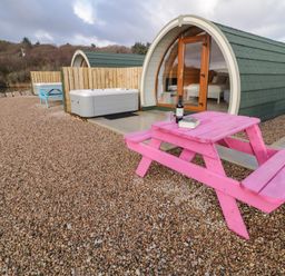 Small curved cabin with glass door next to a pink picnic table and hot tub on a gravel area at The Wheelhouse Pod No 2 in Burtonport County Donegal