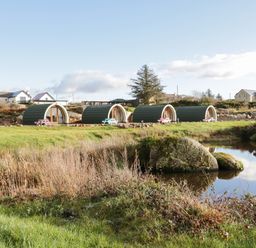 A group of small curved cabins with picnic tables on grass near a pond at The Wheelhouse Pod No 4 in Burtonport County Donegal