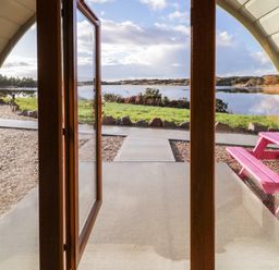 A view through open wooden doors showing a gravel path green grass and a body of water with a pink picnic table outside at The Wheelhouse Pod No 7 Burtonport County Donegal