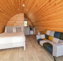 A small room with a bed, colorful sofa, kitchenette, and wooden curved walls at The Wheelhouse Pod No 9 in Burtonport County Donegal
