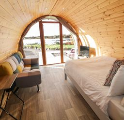 A wooden pod interior with a bed and a multicolored sofa near glass doors opening to a waterside view at The Wheelhouse Pod No 9 in Burtonport County Donegal