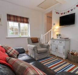 A living room with a leather sofa red throw cushions armchair striped rug small cabinet and wall mounted television at The Little White Cottage in Milton-Under-Wychwood