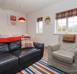 A living room with a black leather sofa and checked armchair near windows with plaid blinds at The Little White Cottage in Milton-Under-Wychwood