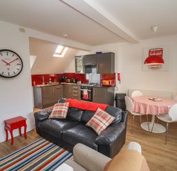 An open plan living room with a black sofa and striped rug beside a kitchen with grey cabinets and red backsplash at The Little White Cottage in Milton-Under-Wychwood