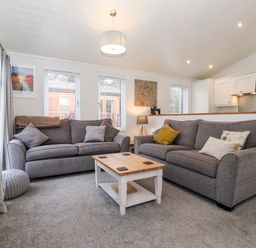 A living room with two gray sofas, a wooden coffee table, windows with blinds, and a view of an open kitchen at Bowness Bay Lodge Oakside 16 Fallbarrow Park near Bowness-On-Windermere