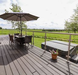 An outdoor deck with a table and chairs under an umbrella and a hot tub overlooking green fields at Holmside in Audley Brow near Market Drayton