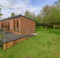 A wooden cabin with glass railing on a deck surrounded by green grass and trees at Holmside Audley Brow near Market Drayton