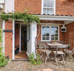 An outdoor seating area with a wooden table and chairs outside a brick cottage with an open door and climbing plants at Beta Cottage in Walberswick