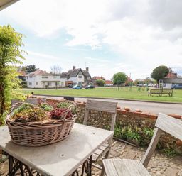 A small outdoor seating area with a table and two chairs overlooking a grassy park with houses and parked cars at Beta Cottage in Walberswick