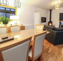 A dining area with a wooden table and chairs and a living room with a gray sofa and a wood burning stove at 2 Strathtay Lodges Aberfeldy