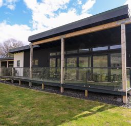 A modern single story house with a wooden porch and glass railing overlooking a grassy yard at 2 Strathtay Lodges in Aberfeldy