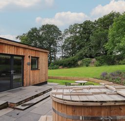 A wooden cabin with glass doors next to a covered wooden hot tub and a garden with grass trees and patio furniture at The Gardener's Retreat in Hutton John near Penruddock