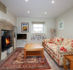 A living room with a floral sofa, wooden coffee table, stone fireplace with a burning fire, wall-mounted TV, window with cushions, and a floor lamp at Tretoil Farm House in Bodmin
