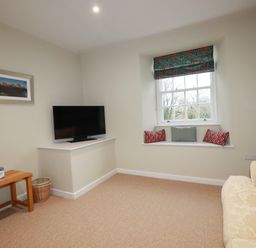 A small sitting room with a window seat holding cushions a wall-mounted television a wooden side table with a tissue box and a sofa at Tretoil Farm House in Bodmin
