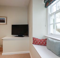 A room corner with a mounted television a wooden side table with a tissue box a wicker basket and a window seat with cushions at Tretoil Farm House in Bodmin