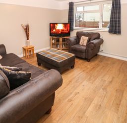 A living room with brown sofas a plaid ottoman a TV showing a fireplace and windows with curtains at Swn Y Mor in Burry Port