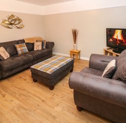 A living room with two sofas a plaid ottoman a television showing a fireplace and wooden flooring at Swn Y Mor in Burry Port