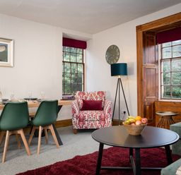 A dining area with green chairs and a wooden table alongside a sitting area with a patterned armchair a round black coffee table and large windows at Watermouth Castle Rhododendron Apartment Berrynarbor near Ilfracombe