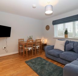 A living room with blue sofa and white cushions a wooden dining table with four chairs a wall mounted television a window with blinds and a potted plant at Ger Y Lli in Rhosneigr