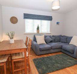 A living room with a wooden dining table and chairs a blue sectional sofa a green rug and a window with striped blinds at Ger Y Lli in Rhosneigr