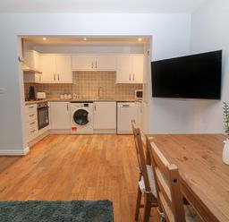 A kitchen area with white cabinets washing machine and dishwasher a wooden dining table with chairs and a wall mounted television at Ger Y Lli in Rhosneigr