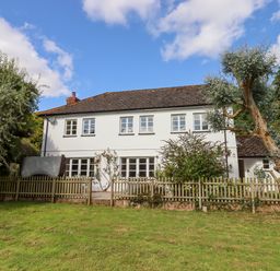 A two story house with white walls and multiple windows behind a wooden fence and trees at Culver Court Cottage in Venny Tedburn near Crediton