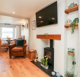 A living room with a leather armchair wooden floor small dining table under a window and a wood stove with a television above at Peppers Cottage in Appledore