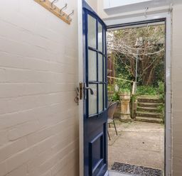 An open blue door leading from a small hallway with white brick walls to an outdoor area with stone steps and garden vegetation at Greenhedges in Budleigh Salterton